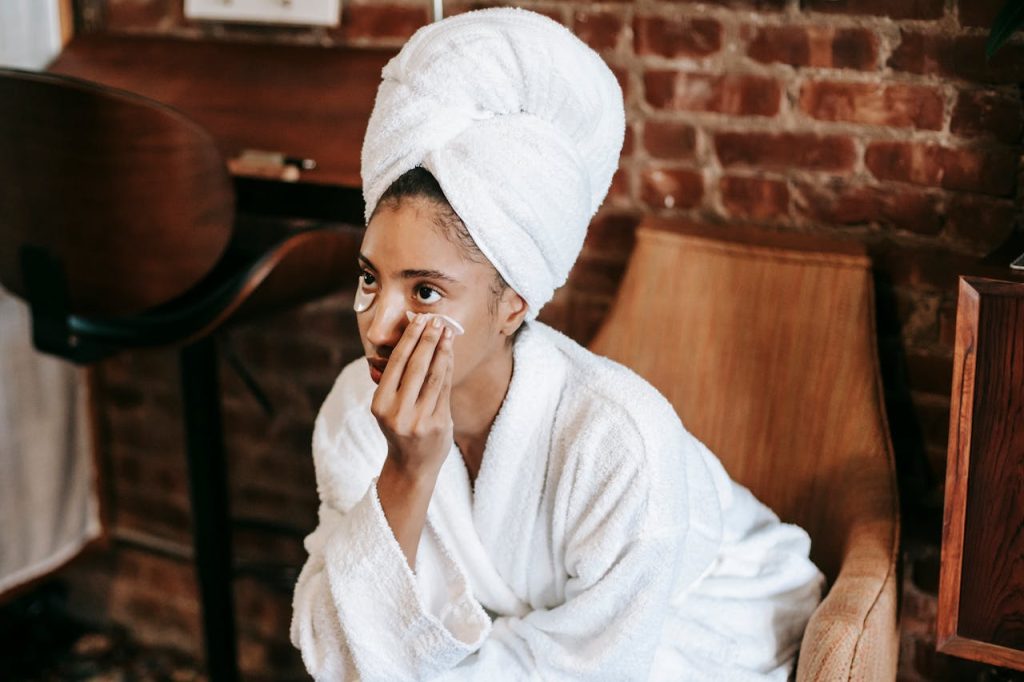 Young female in white bathrobe and towel turban applying eye patches on face while sitting at home